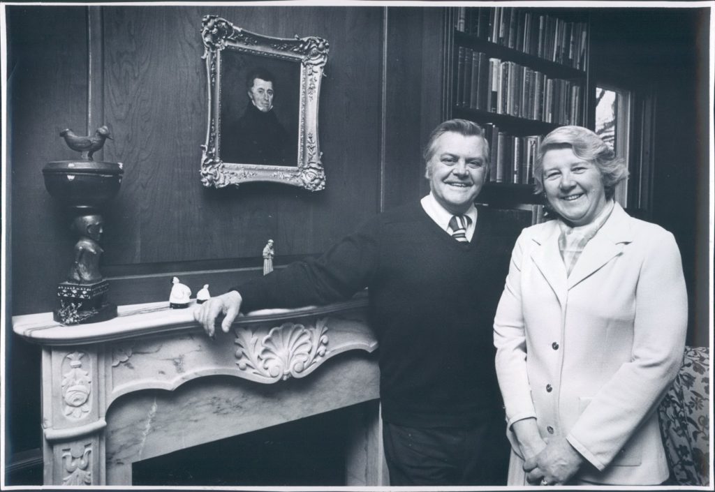 Photo of Elizabeth Brigham and her husband in front of a portrait of her great-grand uncle Ebenezer Brigham.
