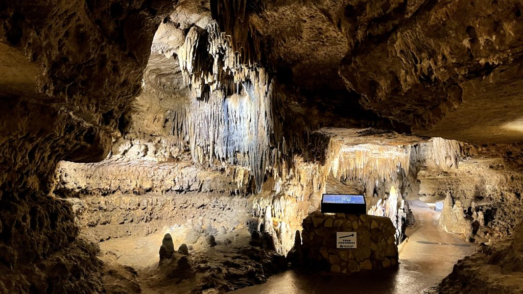 Cathedral Room at Cave of the Mounds with formations coming from the ceiling and on the floor. A light up sign is displayed in the middle with information for guests as they walk through. the caves