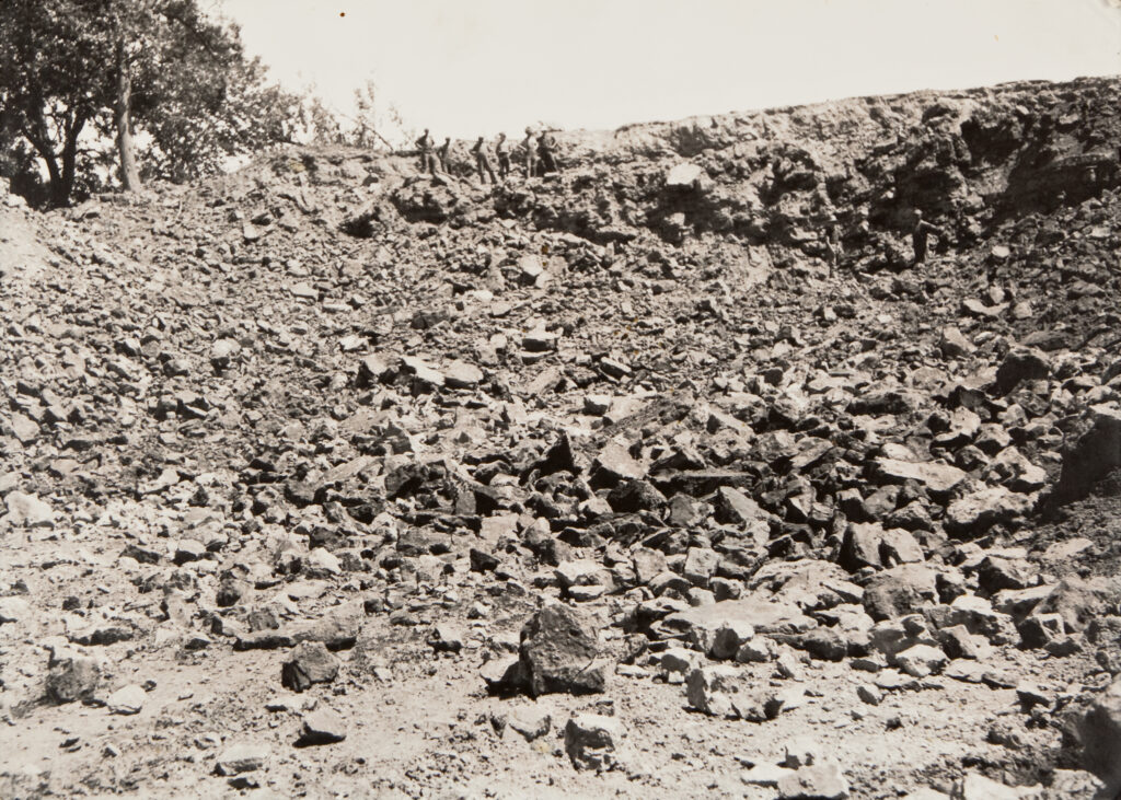 A black and white photo of a large pile of rock and rubble. Several workers gather in a line near the top of the pile. Two holes can be seen near their feet.