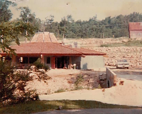A grainy color photo of a large, round building. The hillside is visible in the background.