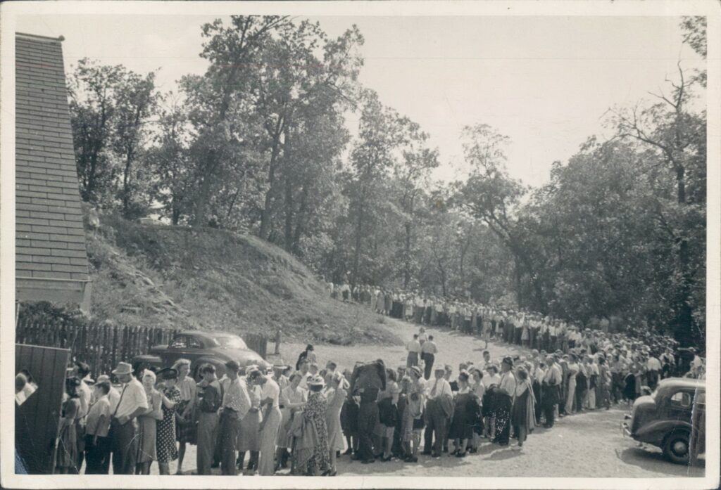 A black and white photo of a large line of several hundred people. The line curves from the cave's entrance up the hill and beyond view.